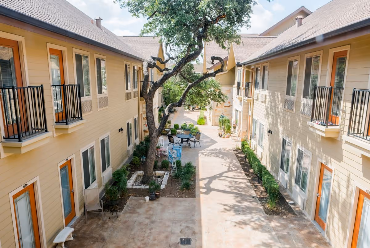 Outdoor courtyard area between two beige two-story buildings with small balconies and orange doors. The courtyard features a large tree in the center, patio tables and chairs, and some potted plants and shrubs along the walkway.
