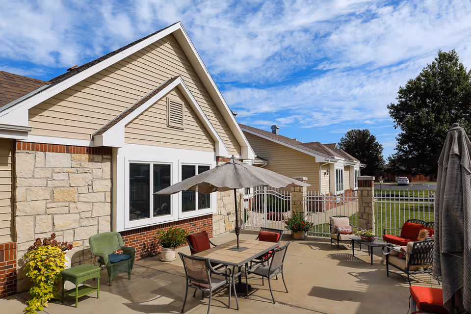 Outdoor patio area at Homestead Assisted & Independent Living of Girard featuring a table with an umbrella, several chairs, potted plants, and a fenced yard with a grassy area and trees under a partly cloudy sky.