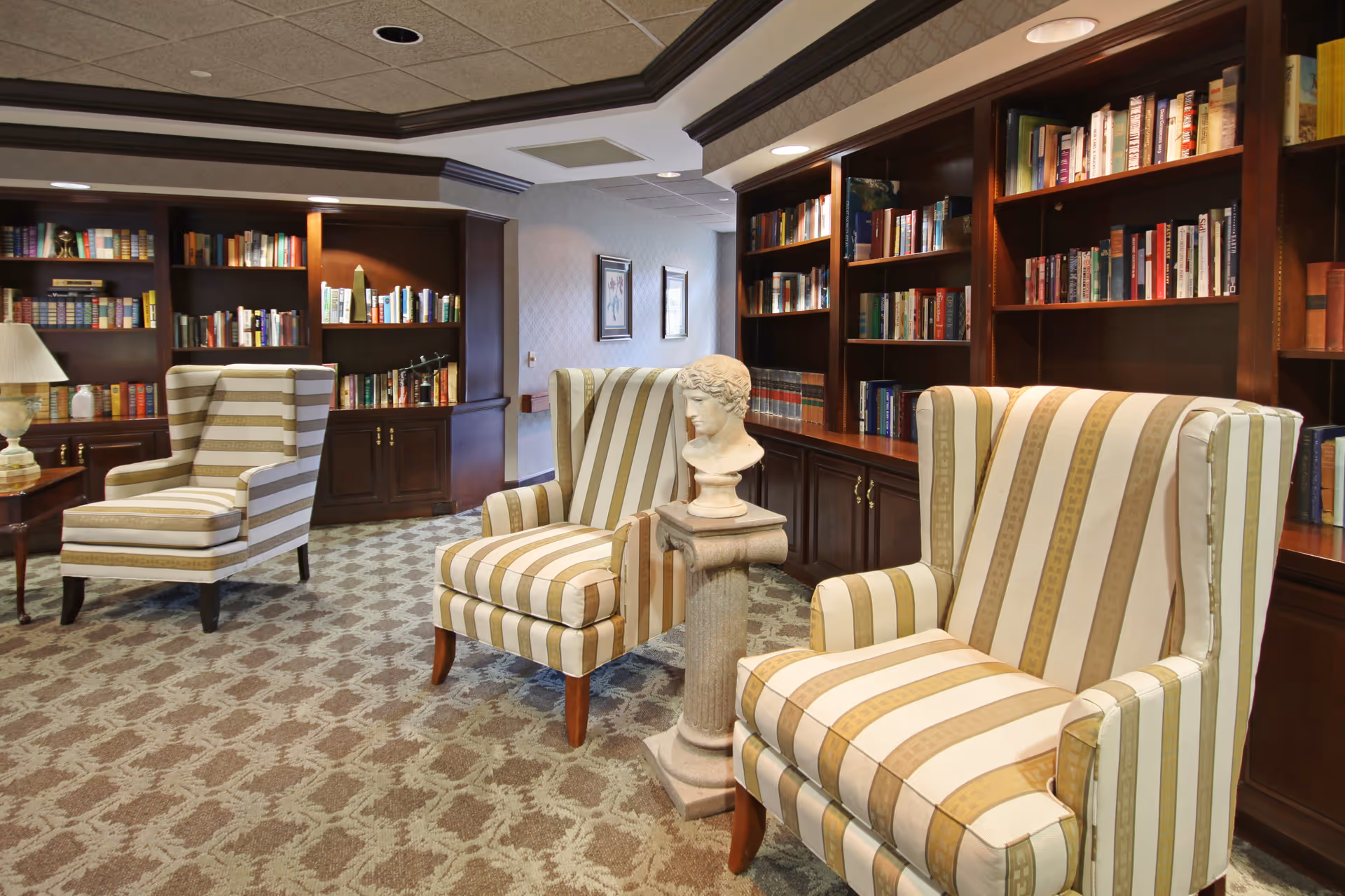 A cozy library-style lounge with striped armchairs, built-in bookshelves and a decorative bust on a pedestal.