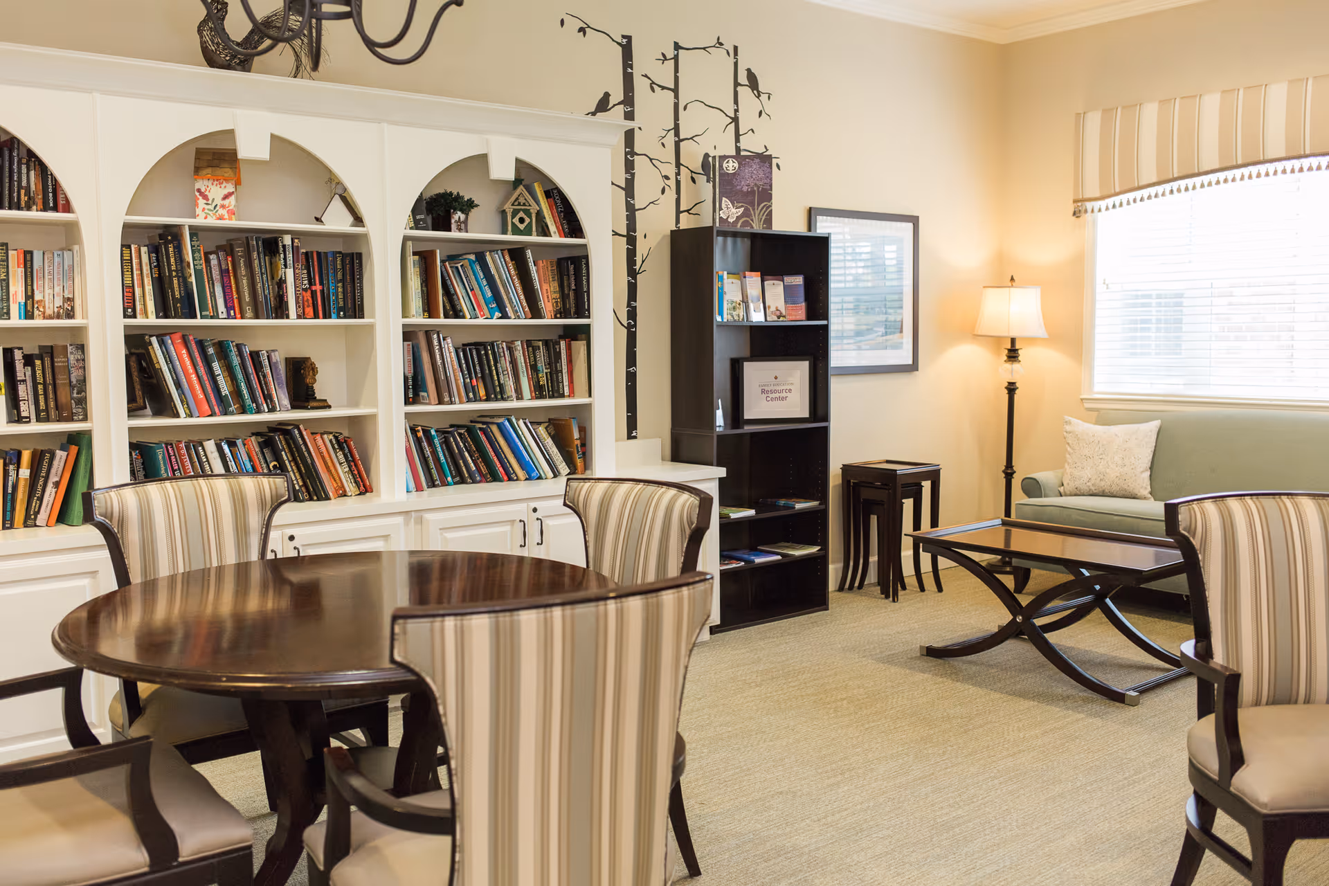 A cozy living room area with a round wooden table surrounded by four striped upholstered chairs, a white built-in bookshelf filled with books, a black bookshelf with pamphlets, a green sofa with a pillow, a glass coffee table, a floor lamp, and a window with blinds and a striped valance.
