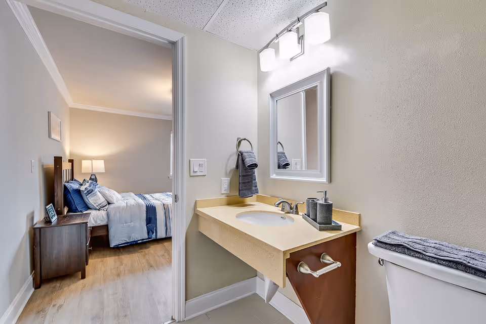 Bathroom vanity with sink, mirror, and towels, opening into a bedroom with a bed and nightstand.