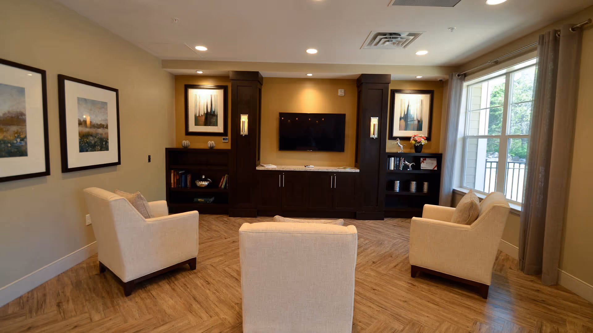 A cozy living room area with three beige armchairs arranged in a semi-circle facing a wall-mounted flat-screen TV. The TV is set within a dark wood built-in cabinet with shelves holding books and decorative items. Two framed artworks hang on the left wall, and a large window with beige curtains lets in natural light on the right side.
