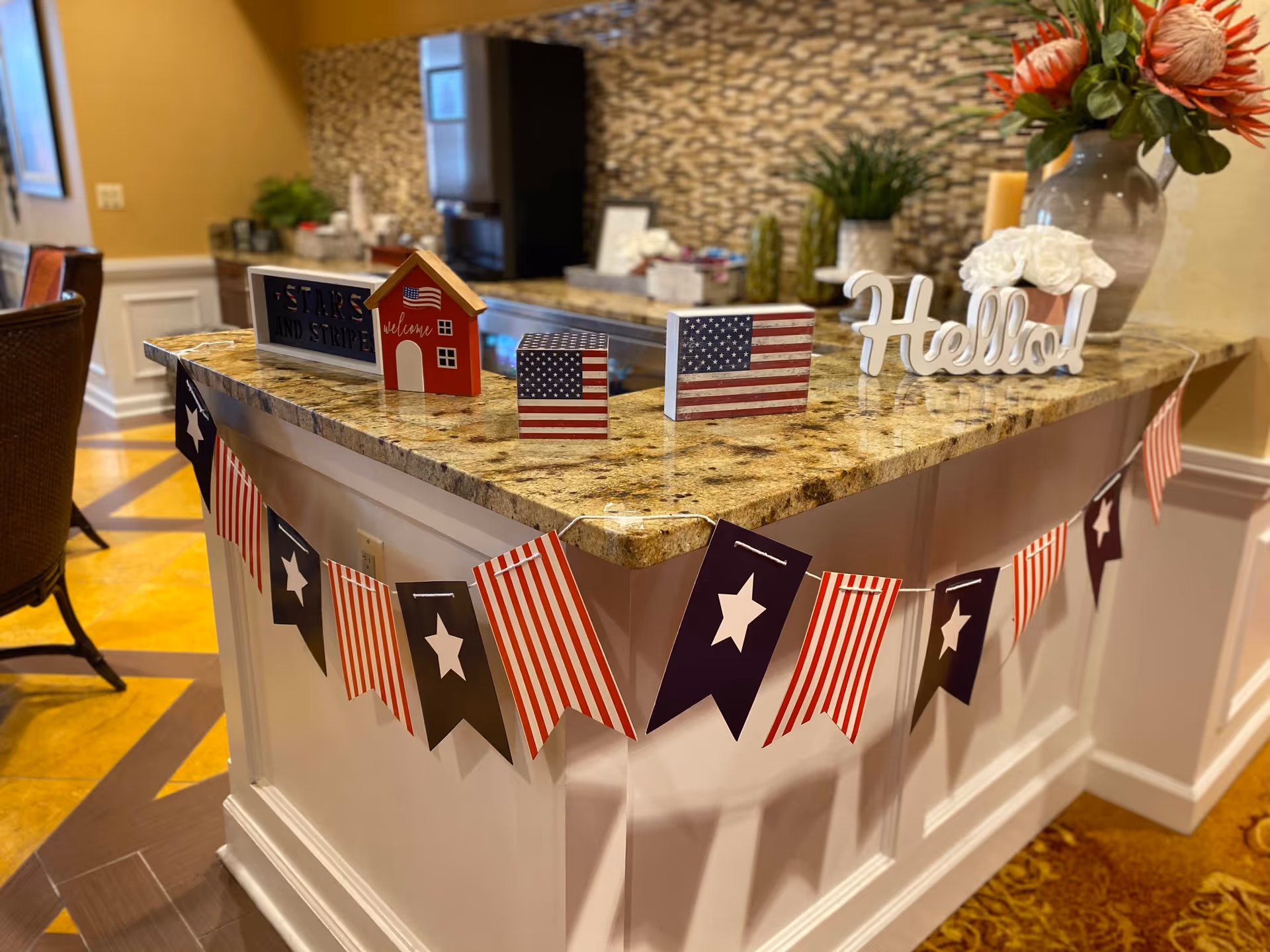 A kitchen or dining area counter decorated with patriotic American-themed items including small wooden blocks painted with the American flag, a small red house decoration with a flag, and a white 'Hello!' sign. A garland with red, white, and blue flags featuring stars and stripes hangs along the front of the counter. In the background, there are plants and a tiled backsplash.
