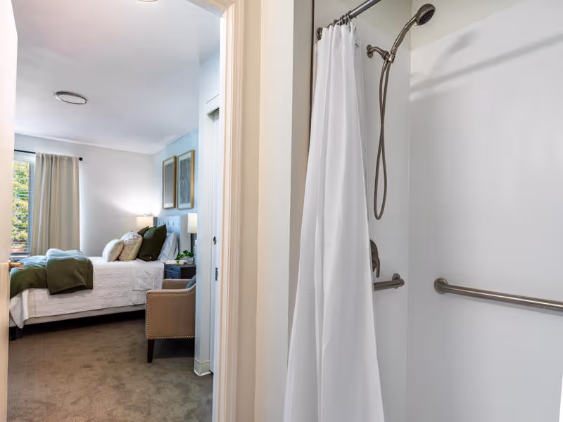 View from a bathroom with a white shower curtain and grab bars into a bedroom with a neatly made bed, green and white bedding, a beige armchair, two framed pictures on the wall, and a window with beige curtains.