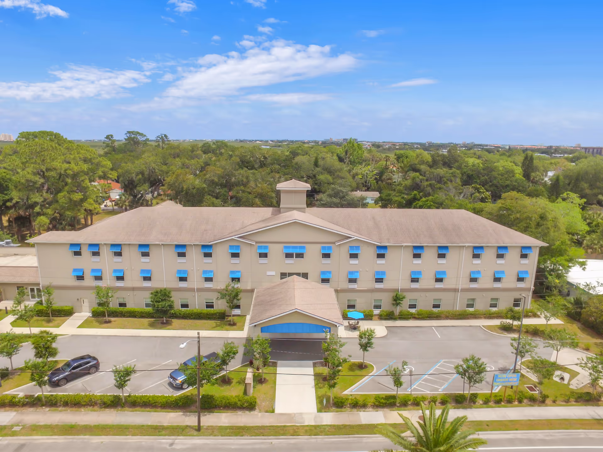 Front exterior view of a two-story senior living facility building with beige walls and blue window awnings, surrounded by trees and a parking lot with a few cars under a partly cloudy sky.