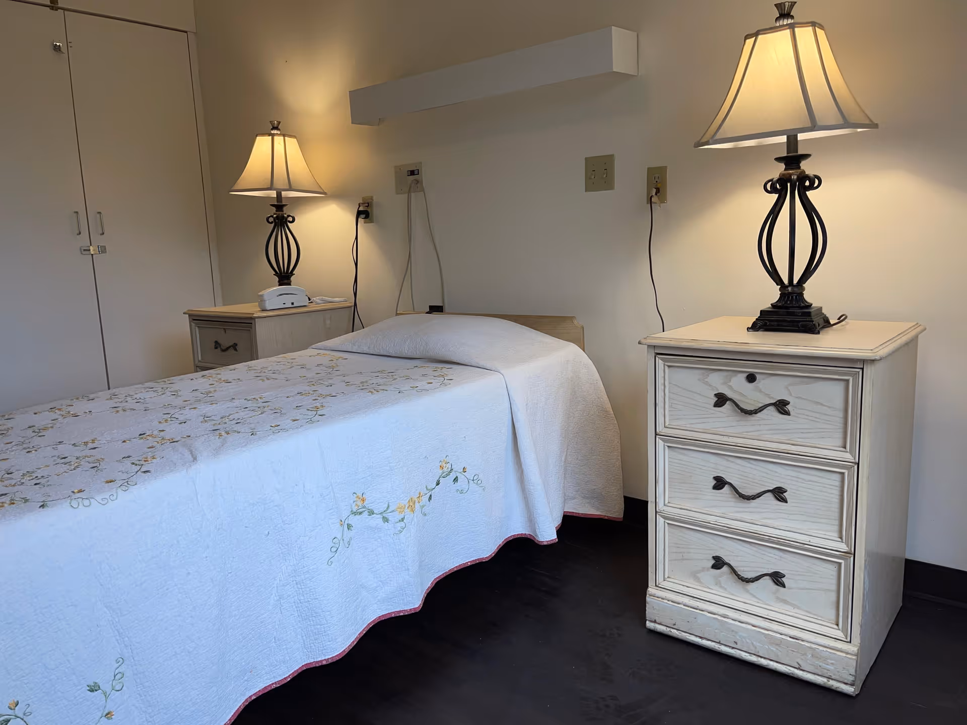 Simple single bed in a small bedroom with a floral bedspread, two bedside tables and lit lamps.