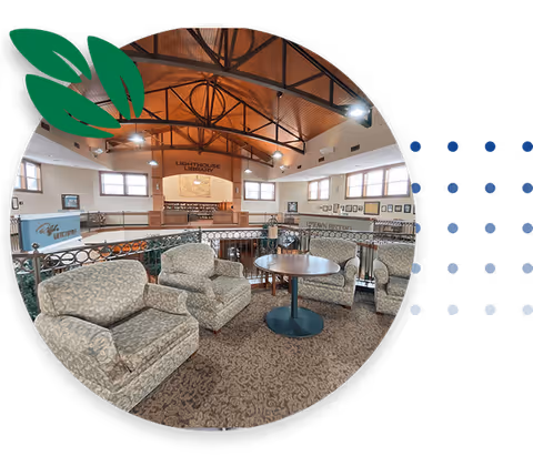 A cozy senior living common area with patterned armchairs arranged around a round wooden table. The space features a high wooden ceiling with exposed beams and large windows letting in natural light. In the background, there is a sign that reads 'Lighthouse Library' above a wooden bookshelf.