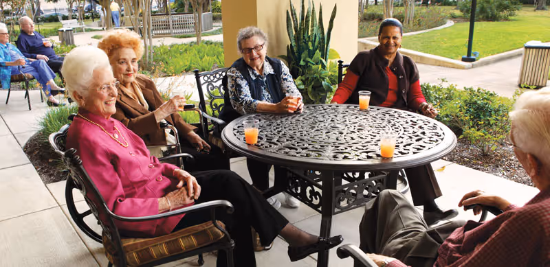 A group of elderly people sitting around a round metal outdoor table with drinks, enjoying a conversation on a patio with greenery and a walkway in the background.