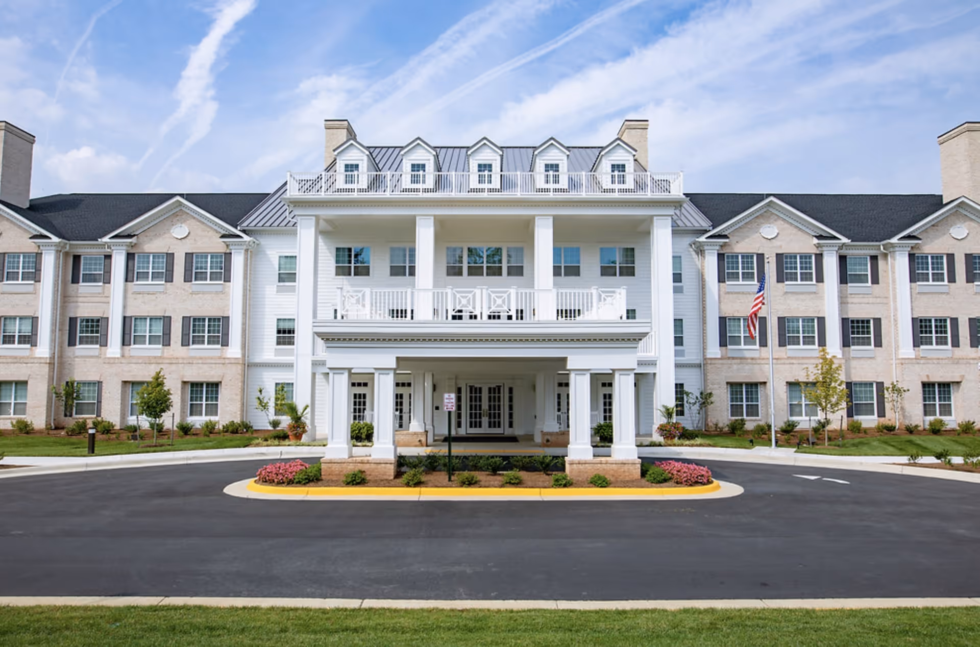 Front entrance and facade of a large three-story senior living building with columns, balconies, and a circular driveway.