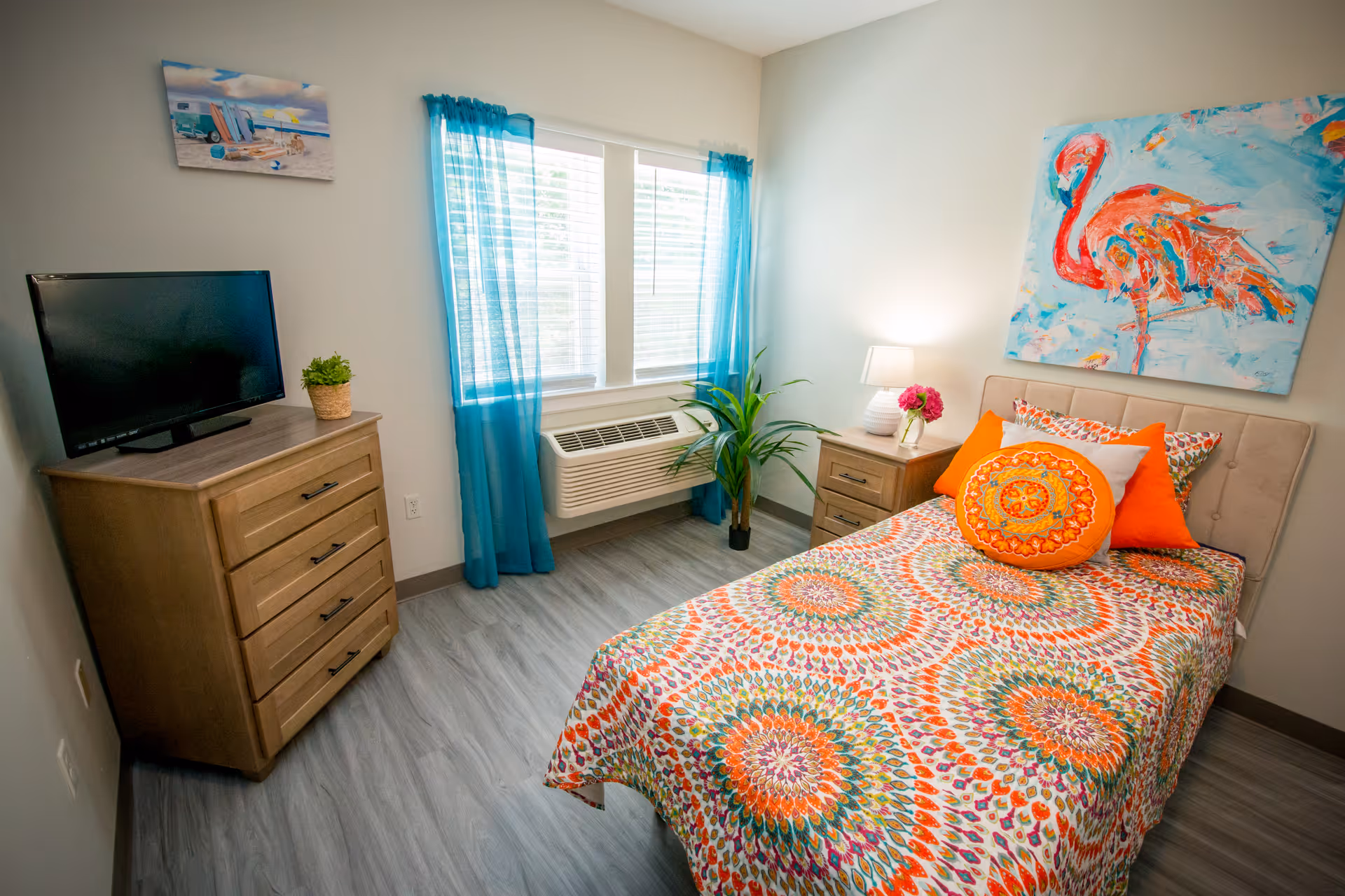 A bright and colorful bedroom featuring a bed with a vibrant patterned bedspread and multiple pillows, a wooden nightstand with a lamp and flowers, a wooden dresser with a TV and a small plant, a window with blue curtains, and a painting of a flamingo on the wall.