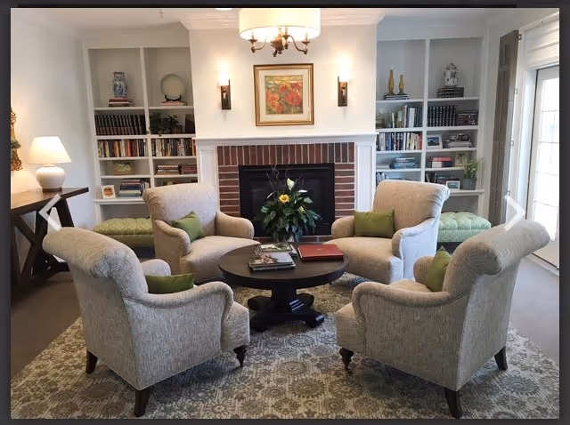 A cozy living room with four beige upholstered armchairs arranged around a round black coffee table. The table has books and a flower arrangement on it. Behind the chairs is a brick fireplace with a floral painting above it, flanked by two wall sconces. Built-in white bookshelves filled with books and decorative items are on either side of the fireplace. A patterned rug covers the floor, and a table lamp is visible on a side table to the left.