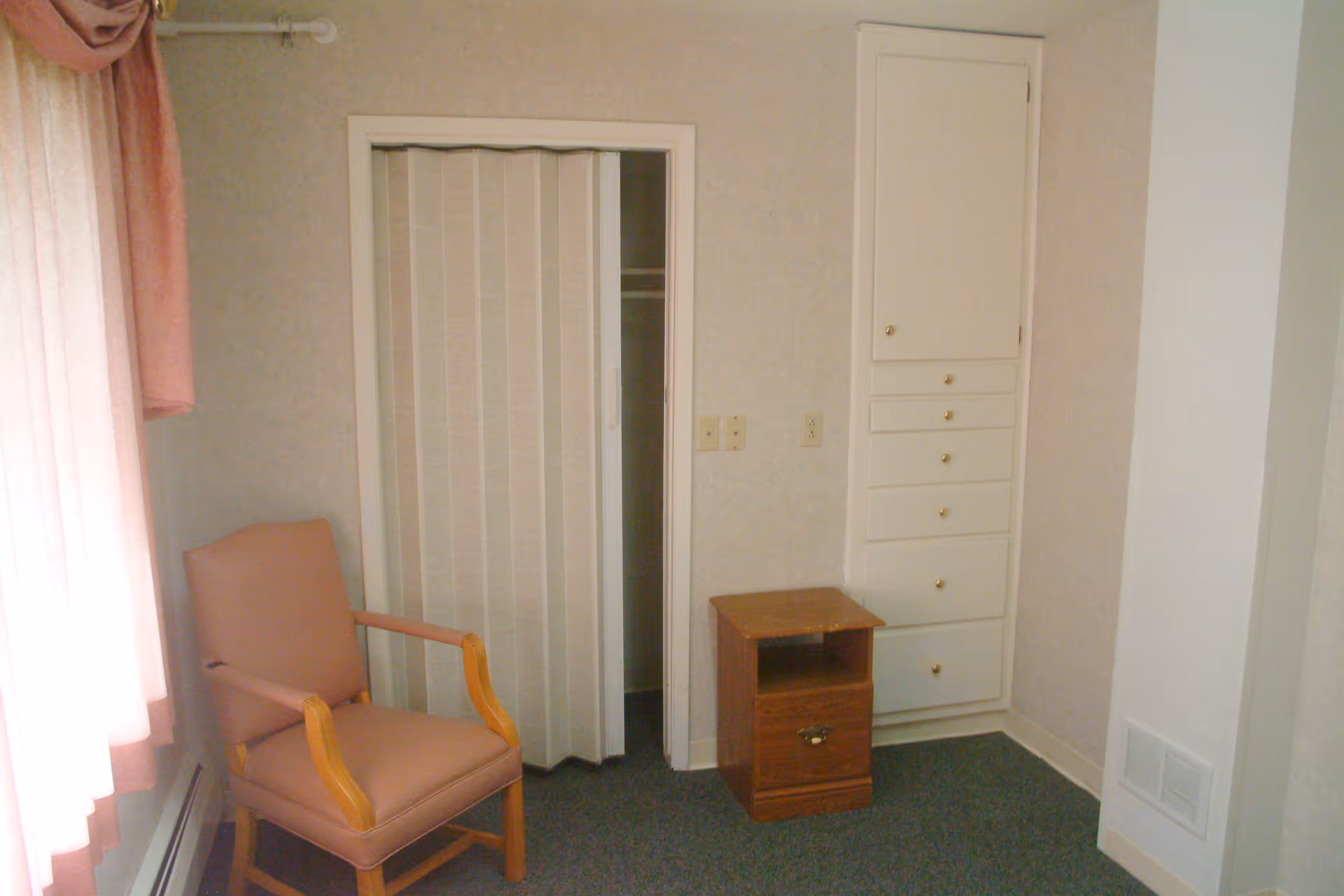 A small room with a pink cushioned wooden armchair near a window with pink curtains, a small wooden nightstand with a drawer, a closet with a white accordion door, and a tall built-in cabinet with multiple drawers and a door.
