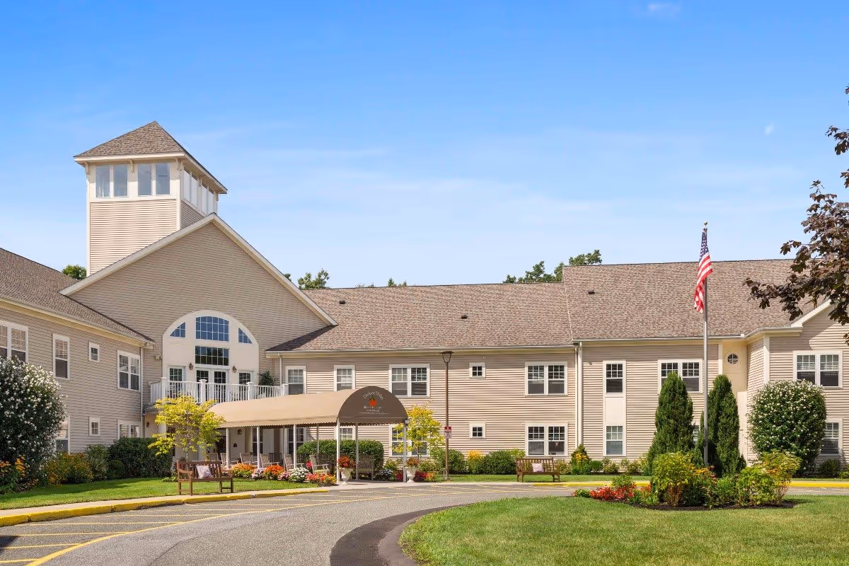 Exterior view of Benchmark at Orchard Valley senior living facility showing a large beige building with multiple windows, a covered entrance with a canopy, landscaped greenery, and an American flag on a flagpole under a clear blue sky.