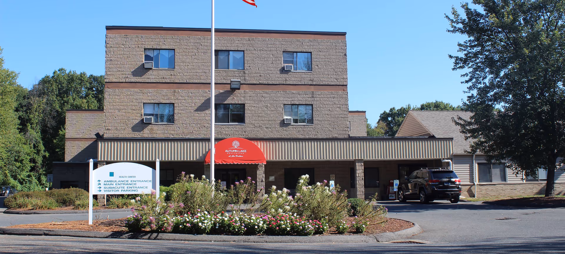 Front exterior view of Autumn Lake Healthcare at New Britain, a three-story building with a red awning over the entrance. There is a flagpole with an American flag in front, surrounded by a landscaped area with flowers and bushes. A sign near the entrance indicates ambulance, main, subacute entrances, and visitor parking. A black SUV is parked near the entrance on the right side.