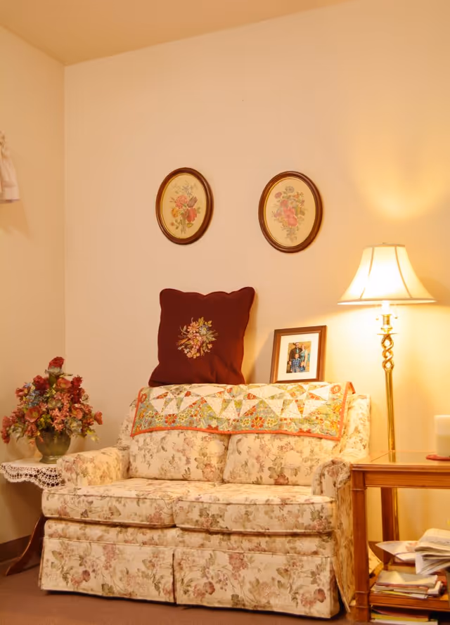 A cozy living room corner with a floral patterned couch adorned with a decorative pillow and a quilt draped over the back. Above the couch are two oval floral paintings. To the right of the couch is a wooden side table with a lamp and a framed photo. To the left is a small table with a lace cover and a vase of flowers.