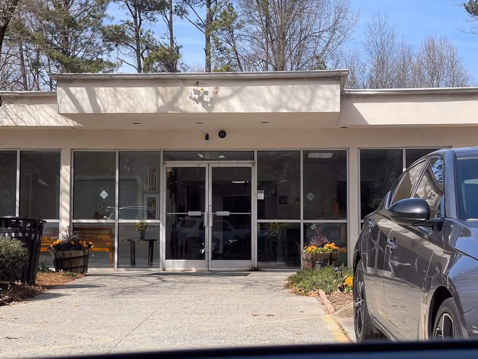 Front entrance of a single-story building with glass double doors, potted flowers, and a car parked at the driveway.