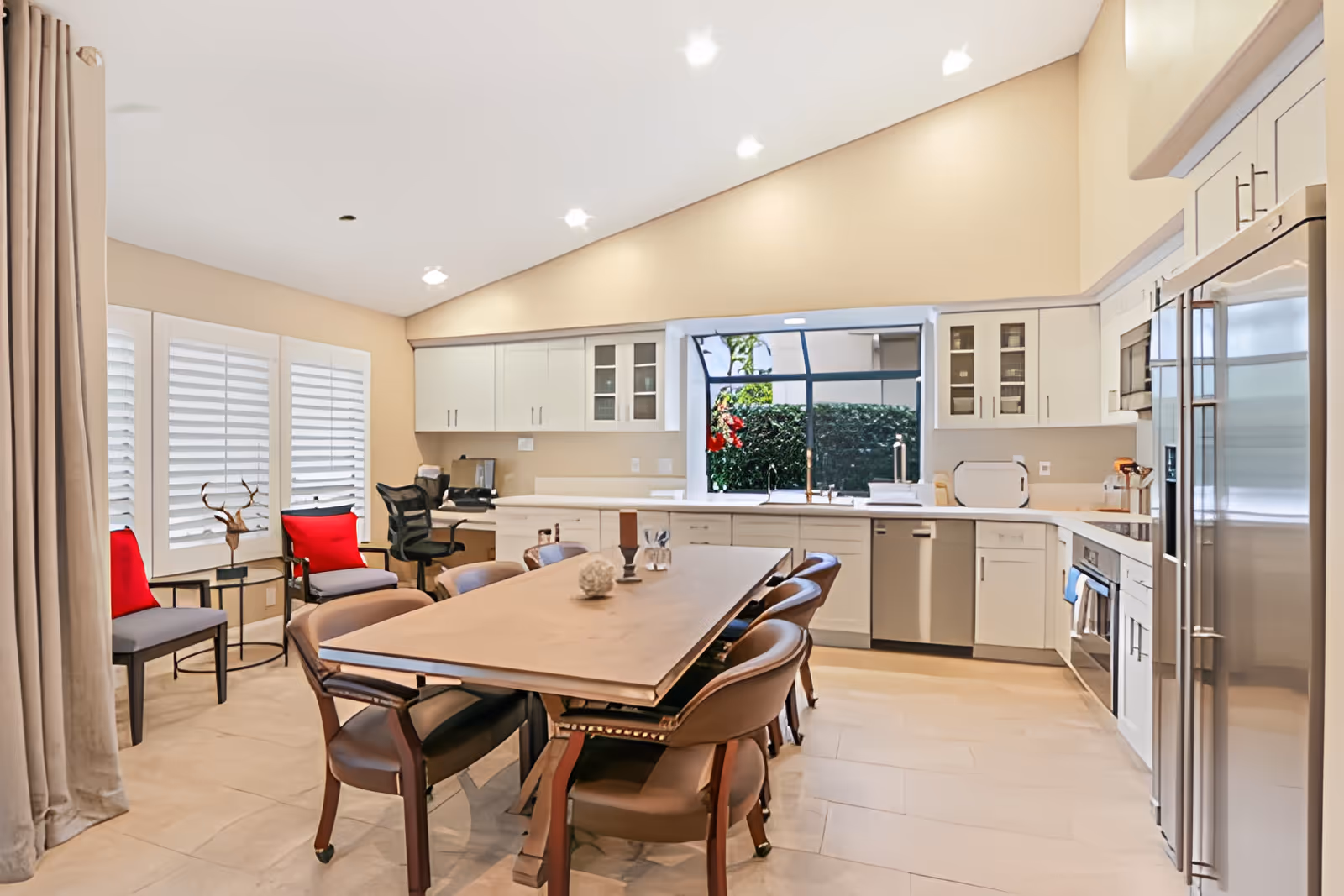 A spacious, modern kitchen with white cabinetry, stainless steel appliances, and a large wooden dining table surrounded by chairs. There are two chairs with red cushions near a window with white shutters, and a desk with an office chair in the corner. A large window above the sink shows greenery outside.