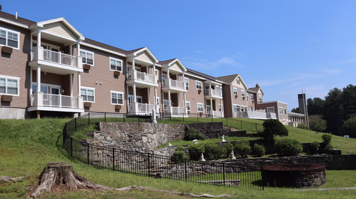 Exterior view of a multi-story residential building with beige siding, white balconies, and multiple windows. The building is situated on a grassy hill with stone retaining walls and black metal fencing. The sky is clear and blue.