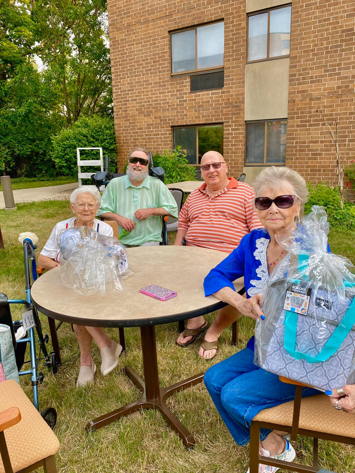 Four elderly individuals sitting around a round outdoor table on grass in front of a brick building. One woman in a blue top and sunglasses is holding a gift basket wrapped in clear plastic. Another woman with white hair and glasses is seated with a walker nearby. Two men, one in a green shirt and sunglasses and the other in a red striped shirt, are also seated at the table. Trees and greenery are visible in the background.