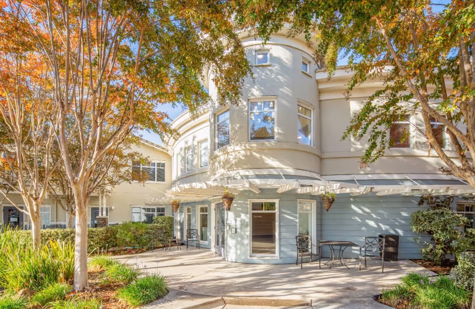 Outdoor patio area of a senior living facility with trees showing autumn foliage, a curved building facade with multiple windows, and outdoor seating including tables and chairs under a pergola.