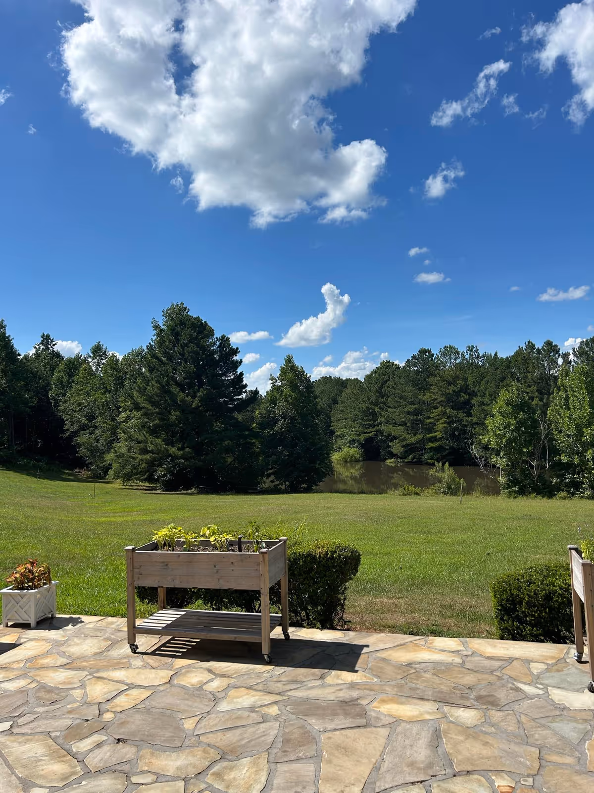 Stone patio with planter boxes overlooking a grassy lawn, trees and a small pond under a blue sky with clouds.