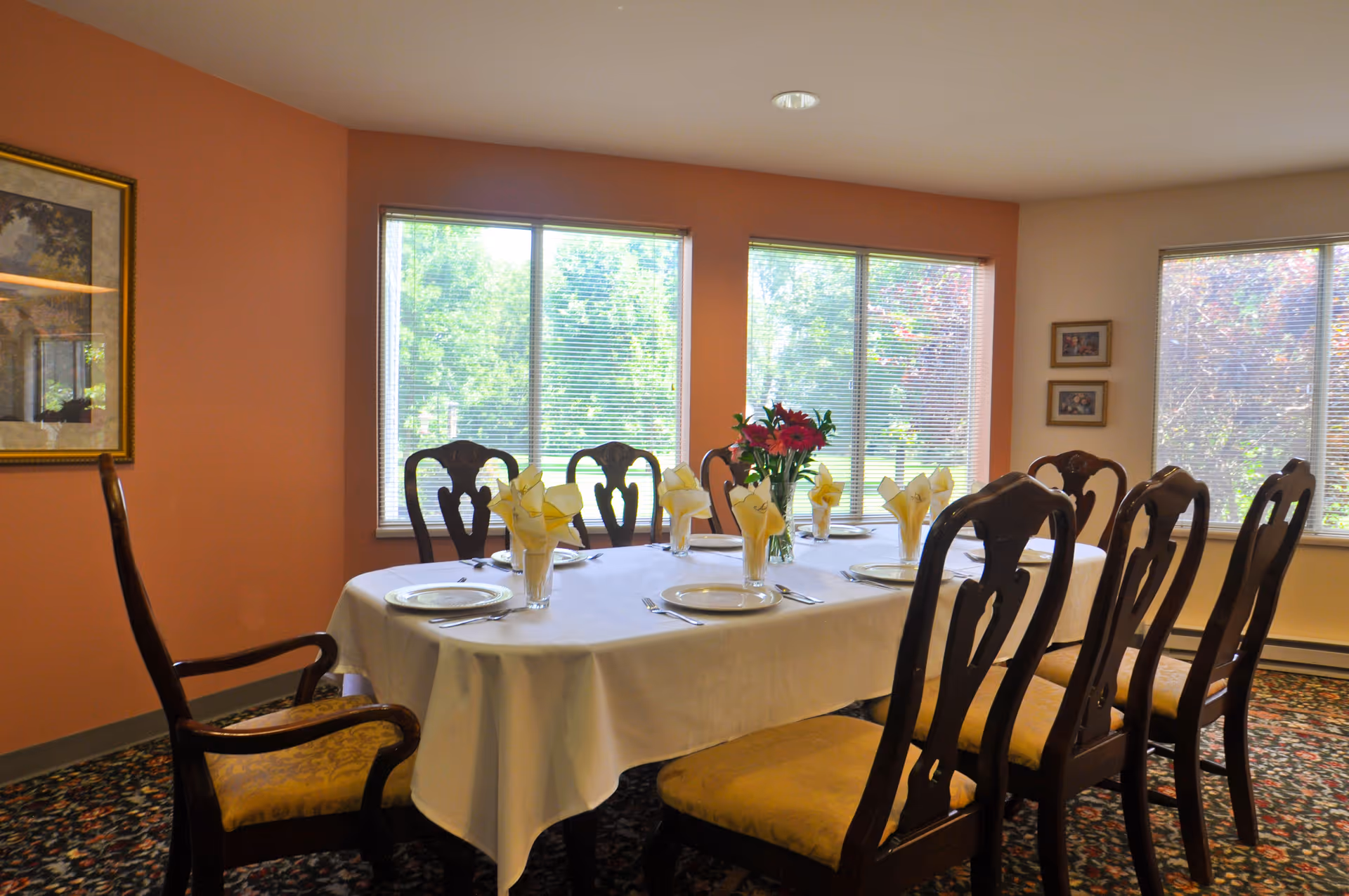 A dining room with a rectangular table covered with a white tablecloth, set with plates, glasses with folded yellow napkins, and silverware. There is a vase with red flowers in the center of the table. The room has large windows with blinds, peach-colored walls, framed artwork, and patterned carpet. Wooden chairs with yellow cushions surround the table.