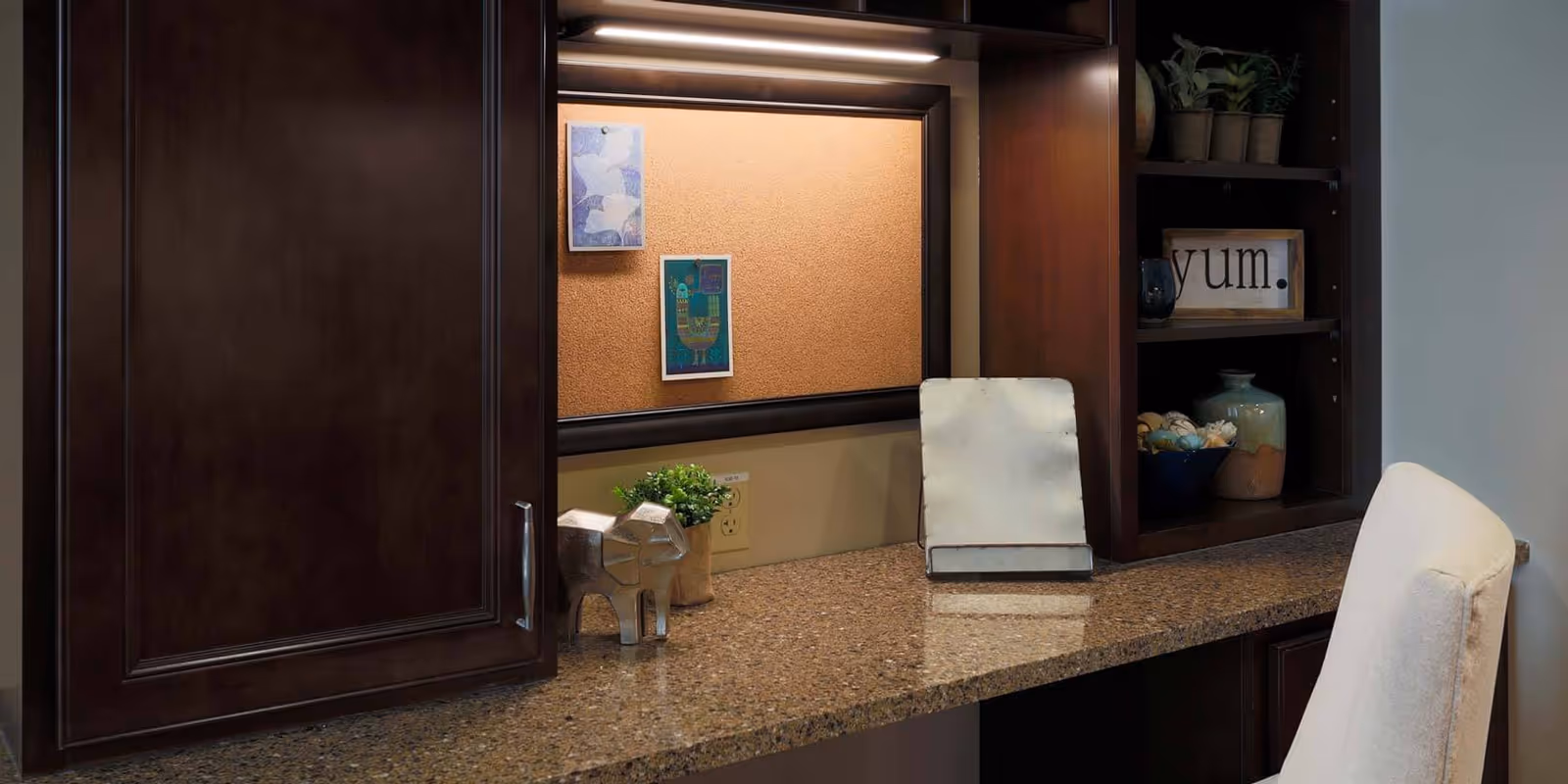 A cozy workspace with a granite countertop, dark wooden cabinets, a cork bulletin board with two pinned pictures, a small potted plant, a silver elephant figurine, and a white cushioned chair. Shelves on the right hold decorative items including a framed sign that says 'yum.' and some pottery.