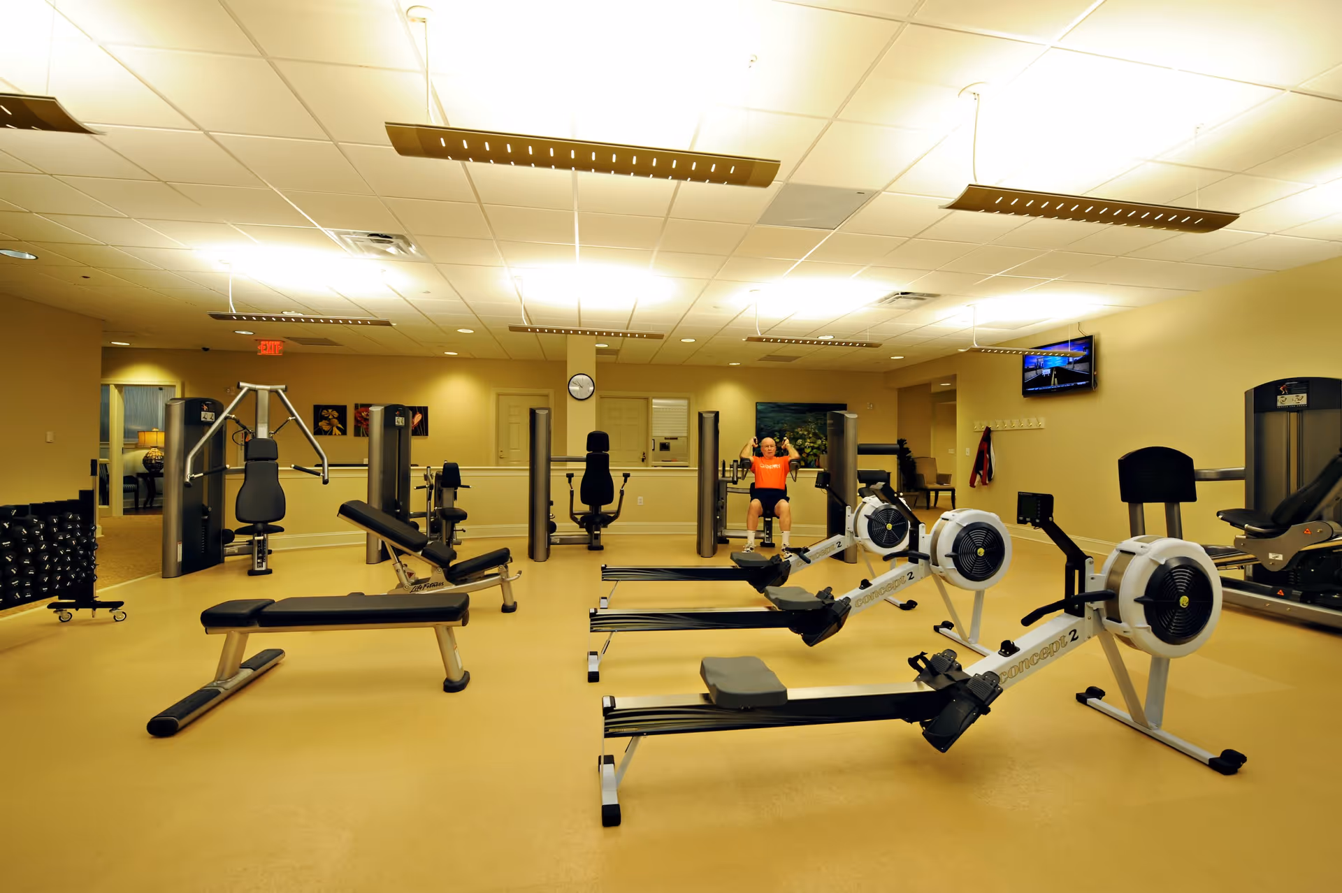 A well-lit fitness room with various exercise equipment including rowing machines, weight benches, and strength training machines. A person wearing an orange shirt is exercising on a seated machine in the background. The room has beige walls, a clock on the wall, and a TV mounted on the right side.