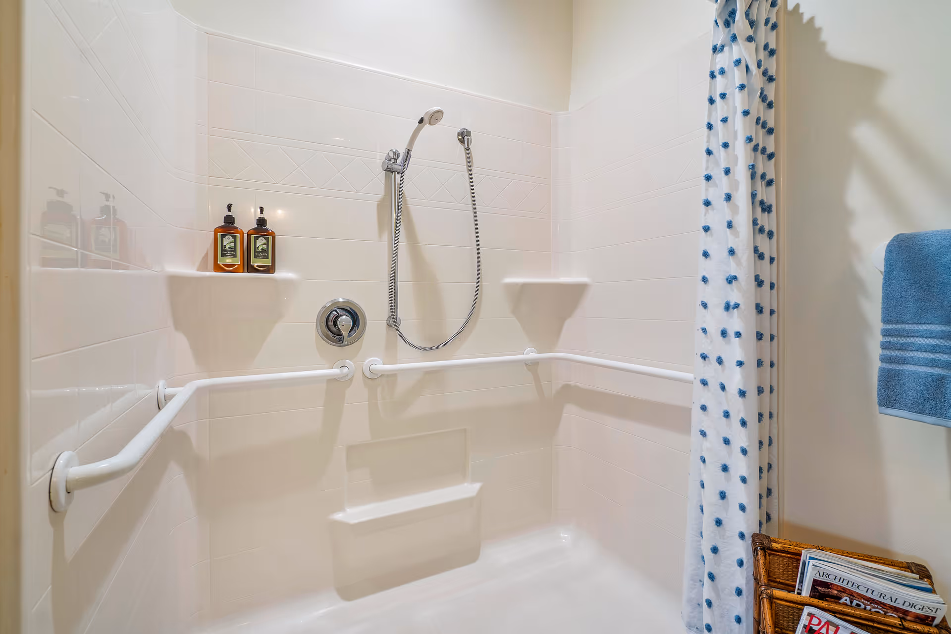 A clean, white tiled shower area with a handheld showerhead, two built-in shelves holding soap dispensers, and white grab bars installed along the walls. A blue and white polka dot shower curtain is partially visible on the right, along with a blue towel hanging on a towel rack and a wicker basket holding magazines.