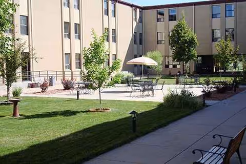 Sunny outdoor courtyard with tables, chairs, a central umbrella, benches, and trees beside a building.
