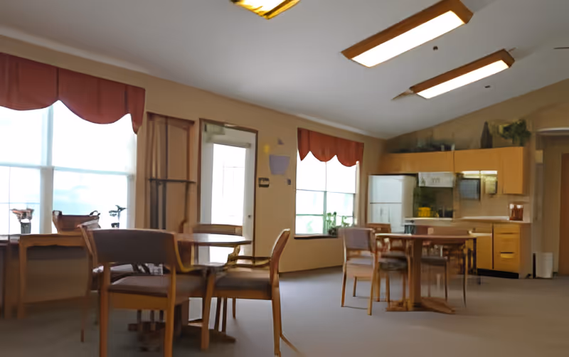 Bright communal dining area with tables and chairs near large windows and a small kitchenette in the background.