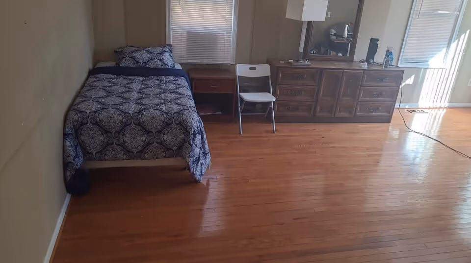 Bedroom in a senior facility with a bed, dresser with mirror, chair, and hardwood floor.