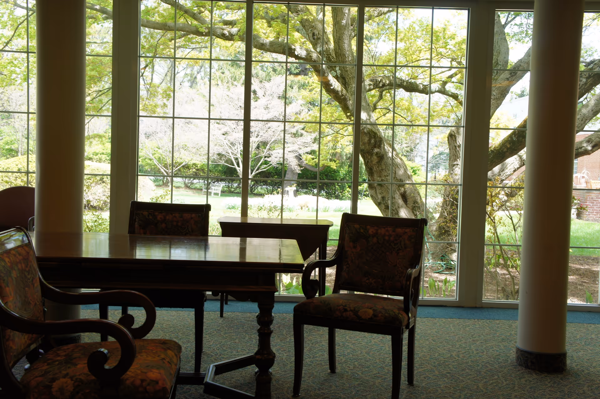 Table and upholstered chairs in a common room with large multi-pane windows showing trees and a garden outside.