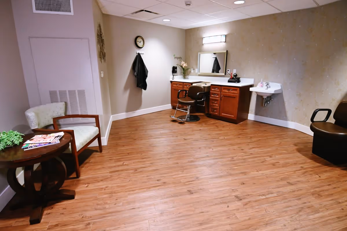 A room with wooden flooring featuring a salon chair in front of a counter with drawers and a mirror, a small wall-mounted sink, two chairs, and a small round table with magazines and a plant on it.
