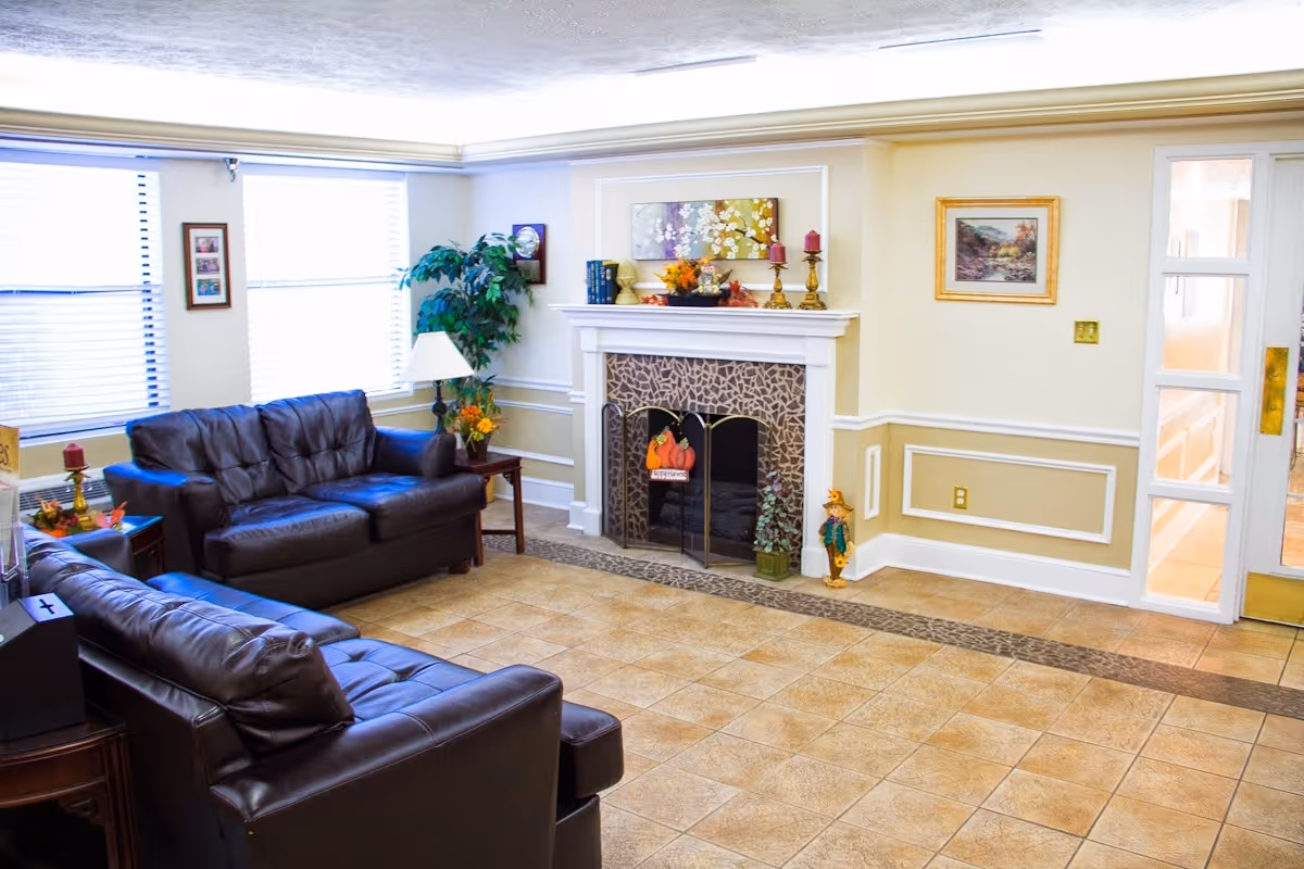 A bright living room with two dark brown leather sofas facing a white fireplace decorated with autumn-themed items and candles. The room has tiled floors, beige walls with white trim, a large window with blinds, a potted plant, framed pictures on the walls, and a glass door on the right side.
