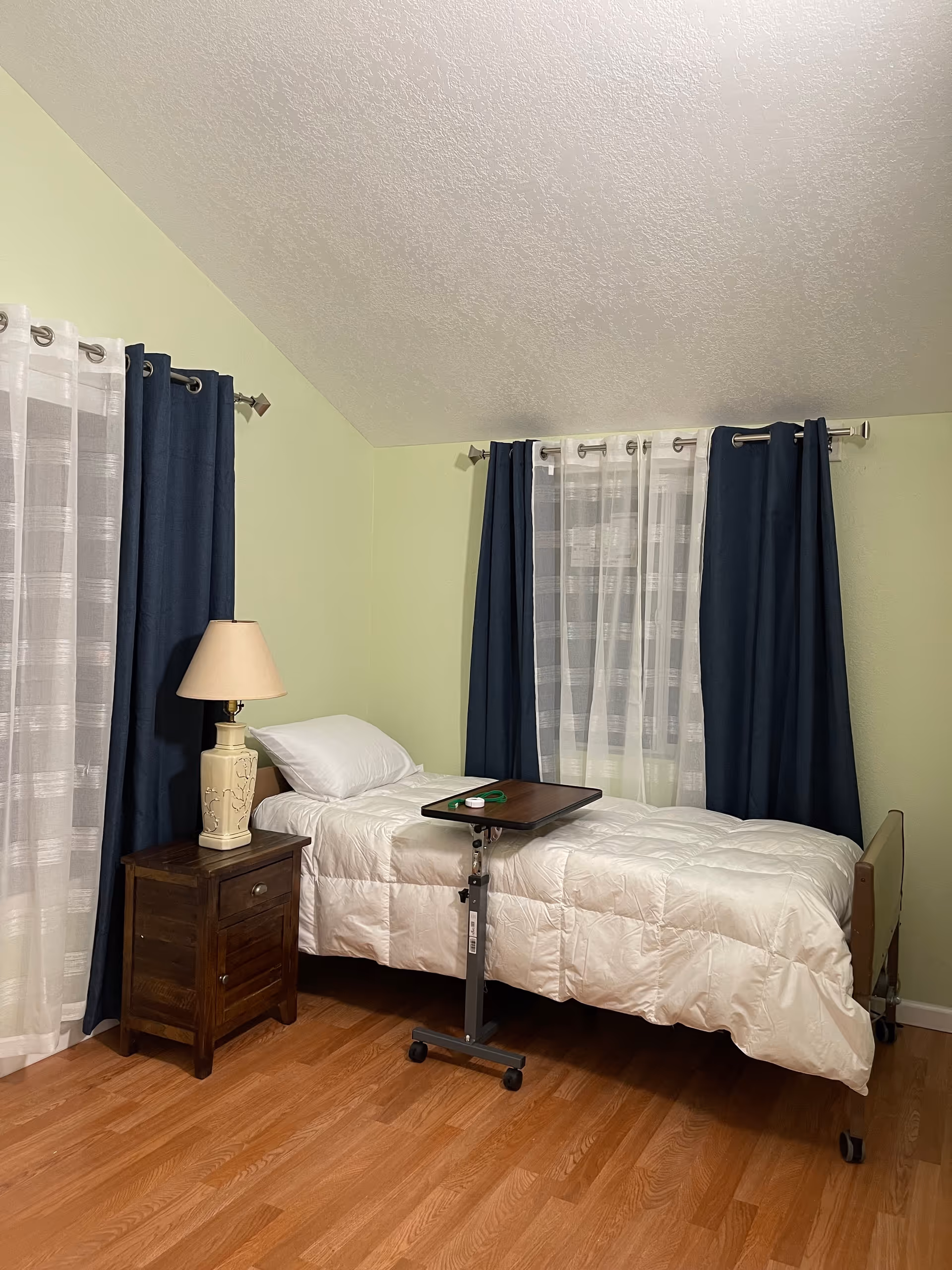 A small bedroom with a single bed covered in a white comforter and a white pillow. Next to the bed is a wooden nightstand with a beige lamp on it. There is a small adjustable overbed table on wheels positioned over the bed. The room has light green walls, wood flooring, and two windows with white sheer curtains and dark blue drapes.