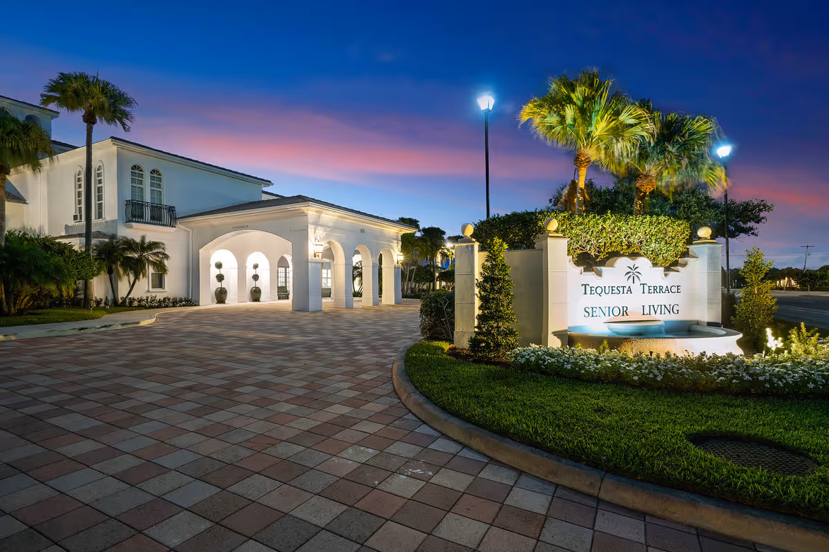 The illuminated entrance and sign for Tequesta Terrace Senior Living in front of a Mediterranean-style building at dusk.
