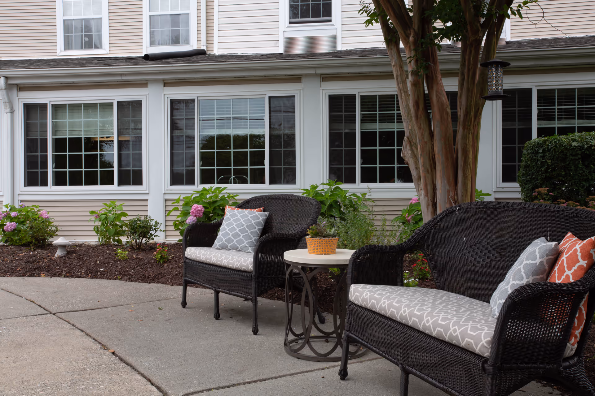 Outdoor seating area with two black wicker chairs and a matching loveseat with patterned cushions and pillows, a small round table with a potted plant, situated on a concrete patio in front of a building with large windows and beige siding, surrounded by green plants and flowers.