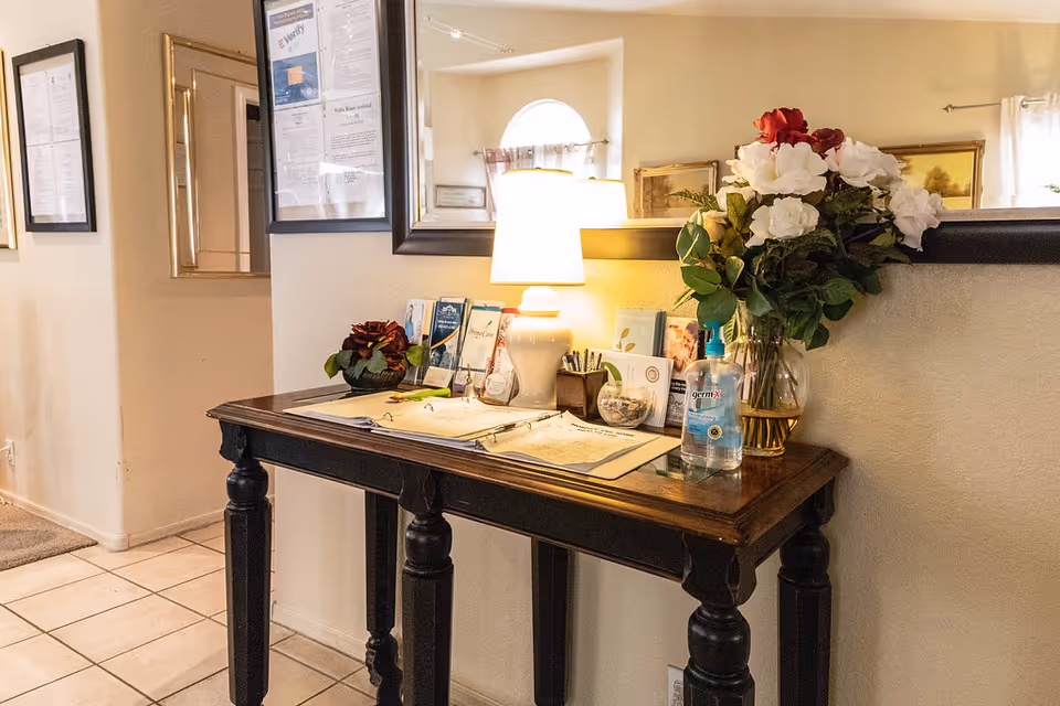 A wooden console table with a dark finish against a beige wall, decorated with a table lamp, a vase of red and white flowers, hand sanitizer, brochures, and a guest sign-in book. Above the table is a large mirror reflecting part of the room, and framed documents are hung on the adjacent wall.