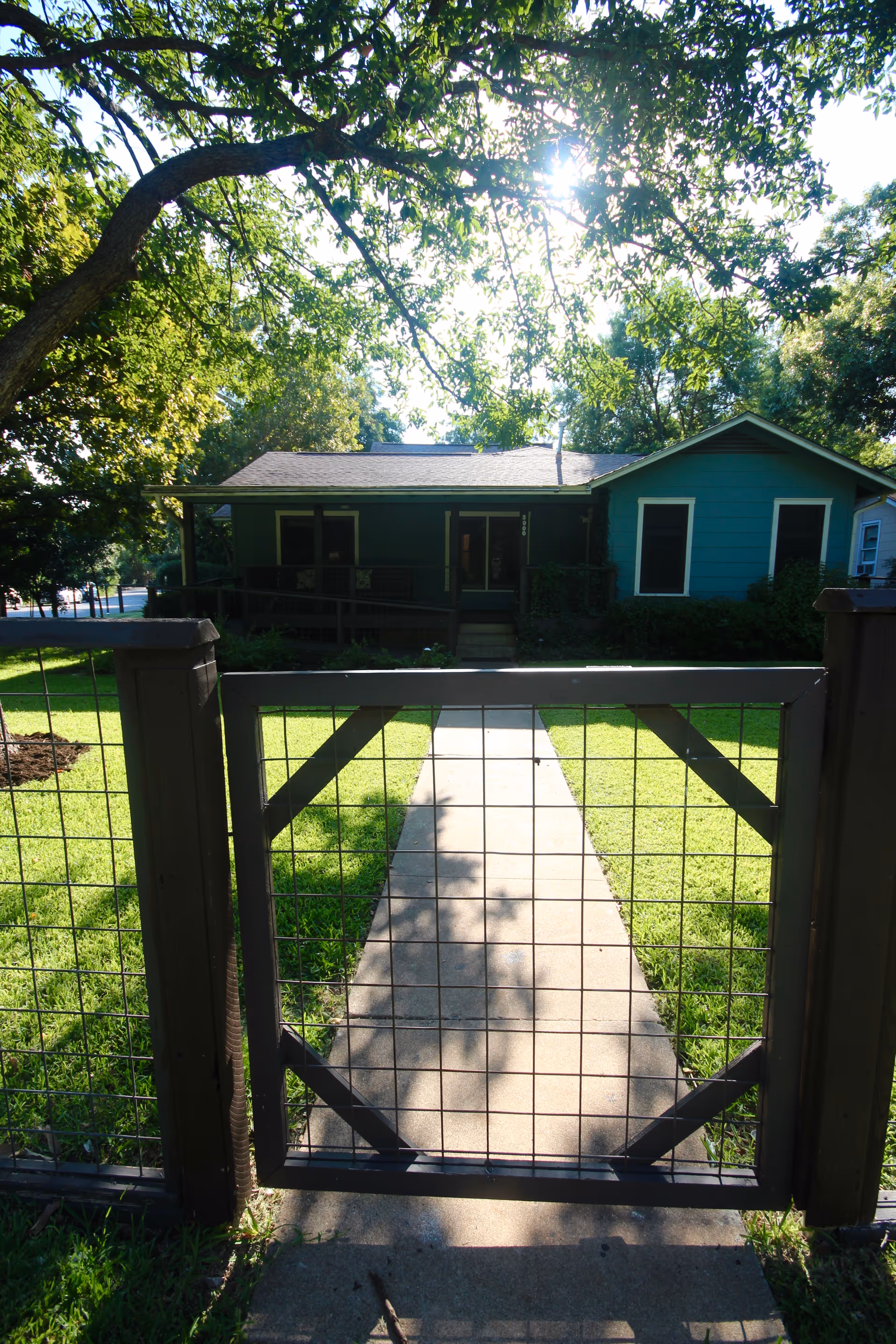 View of a single-story blue house with a front porch, seen through a metal gate and fence. A concrete pathway leads from the gate to the house entrance, surrounded by green grass and shaded by large trees.