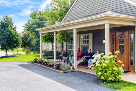 Three elderly people sitting and relaxing on chairs on the covered front porch of a residential building with a wooden door, surrounded by green plants and flowers under a blue sky.