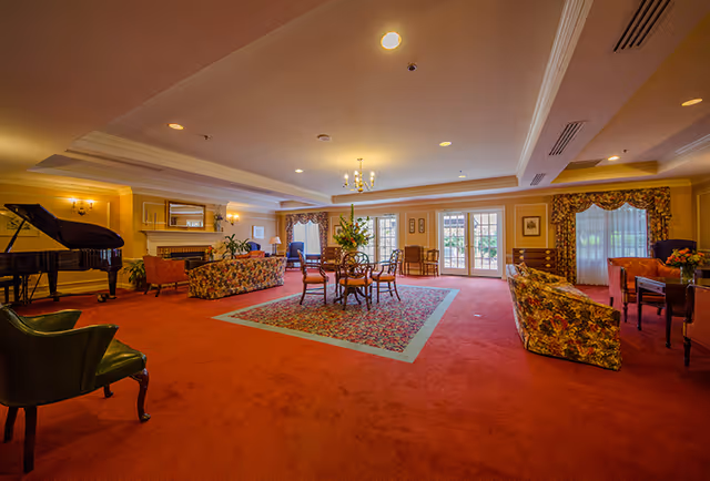 A spacious living room area with red carpet and floral-patterned furniture including sofas and chairs. There is a grand piano on the left side near a fireplace with a mirror above it. A round table with chairs is centered on a floral rug. Large windows and glass doors allow natural light to enter the room, and floral curtains frame the windows. The ceiling has recessed lighting and a chandelier.
