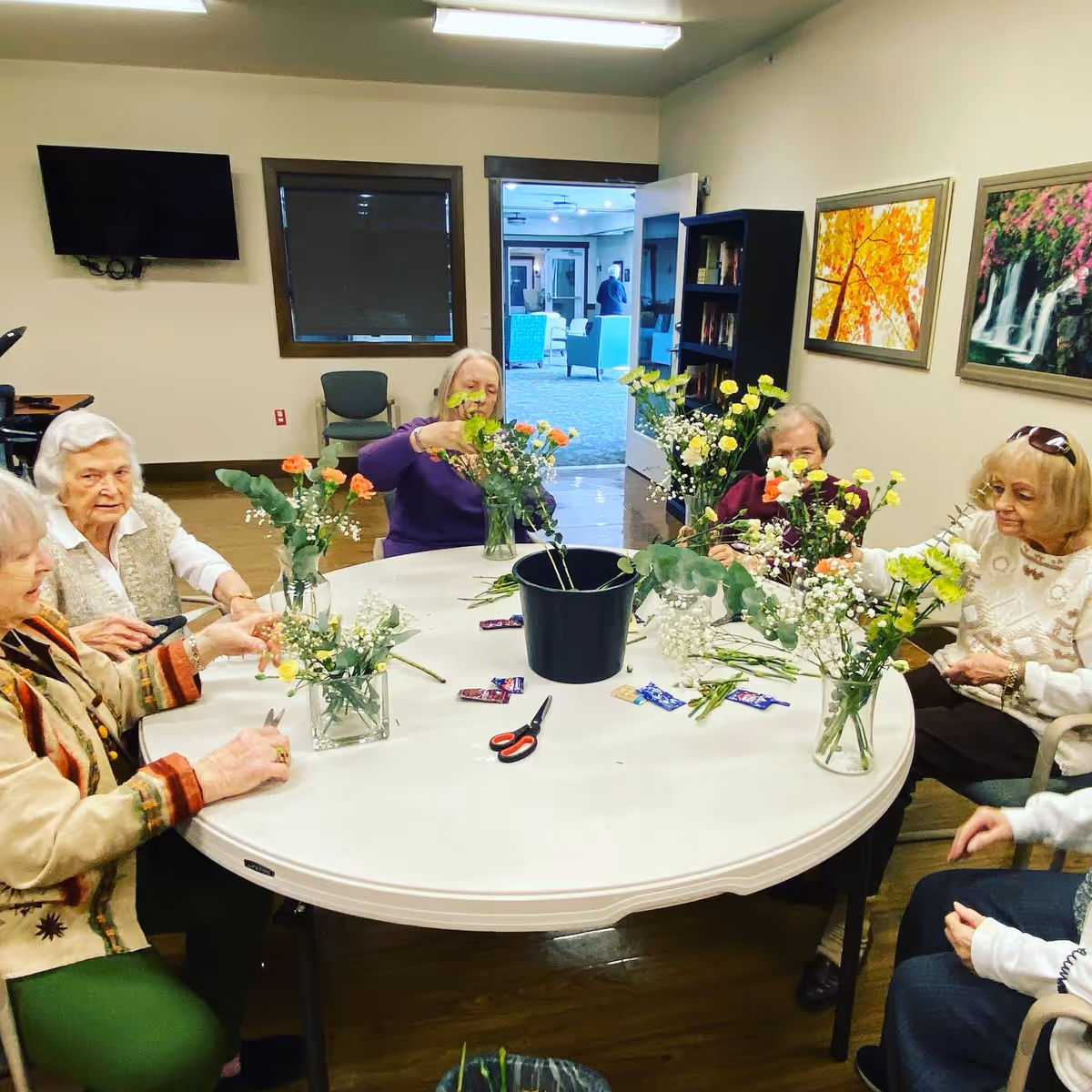 A group of elderly women sitting around a round table in a senior living facility, arranging flowers in vases. The room has a TV mounted on the wall, a bookshelf, and framed pictures of nature scenes. There are scissors and flower stems on the table.