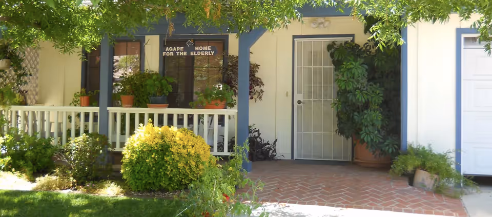 Front porch of Agape Home For The Elderly with a white railing, potted plants on the porch and around the entrance, a white door with a security screen, and a sign above the window that reads 'Agape Home For The Elderly'.