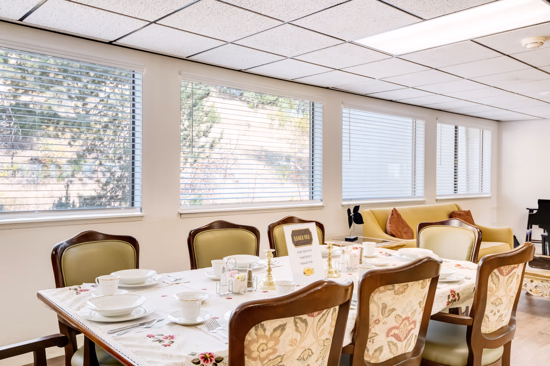 A dining room with a long table set for a meal, featuring white plates, cups, and silverware on a floral tablecloth. The room has several wooden chairs with cushioned seats and backs around the table. Large windows with blinds allow natural light to fill the space. In the background, there is a yellow sofa with two brown pillows and a black piano on the right side.