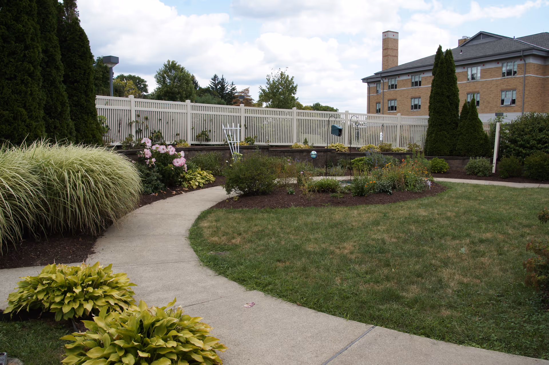 A landscaped outdoor garden area with a curved concrete pathway surrounded by green grass, bushes, and flowering plants. There is a white fence in the background and a multi-story brick building with several windows behind the fence. The sky is partly cloudy.
