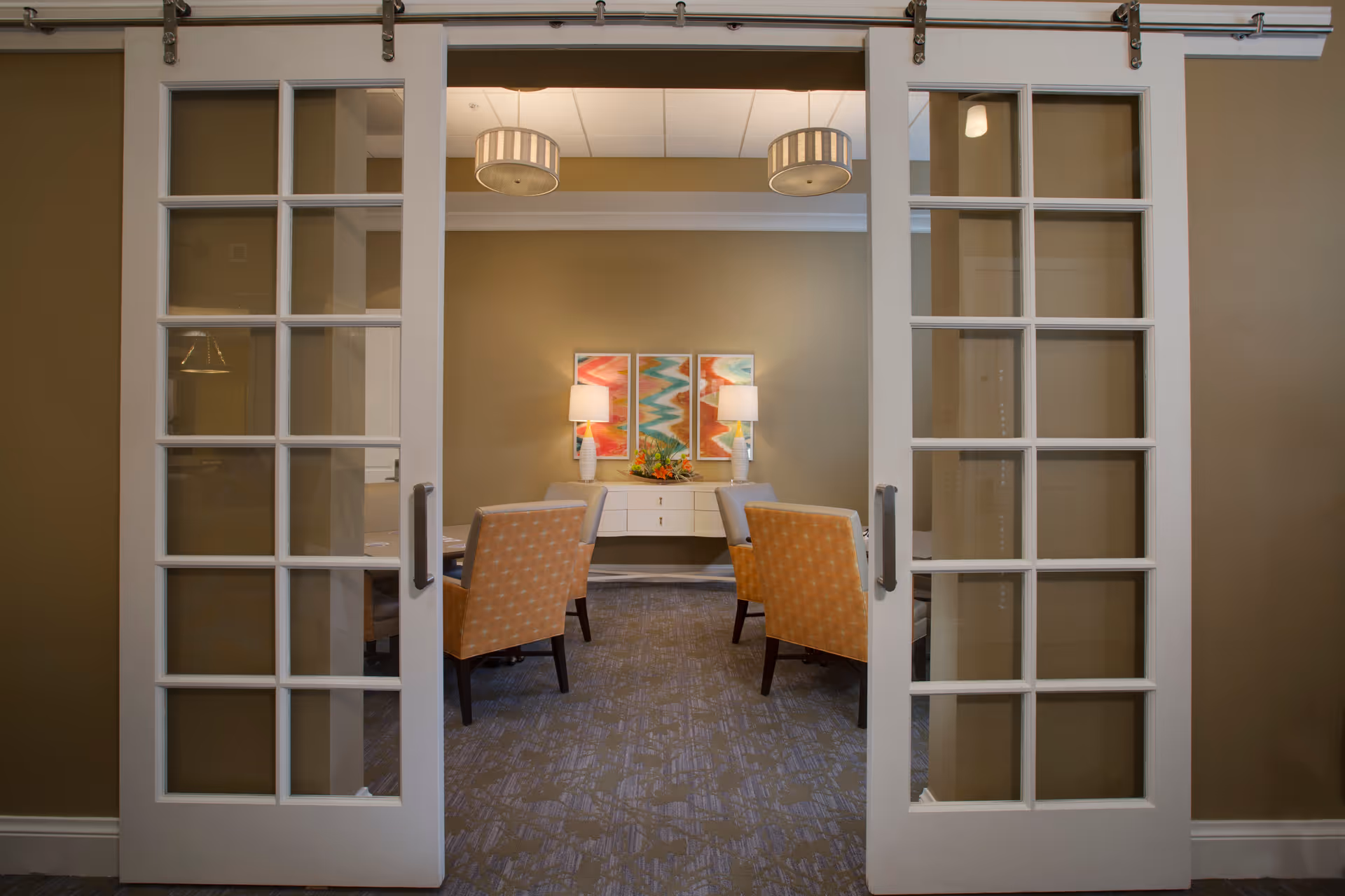 View through white sliding glass-paneled doors into a room with four upholstered chairs around a table, a white sideboard with two lamps, and colorful abstract artwork on the wall.