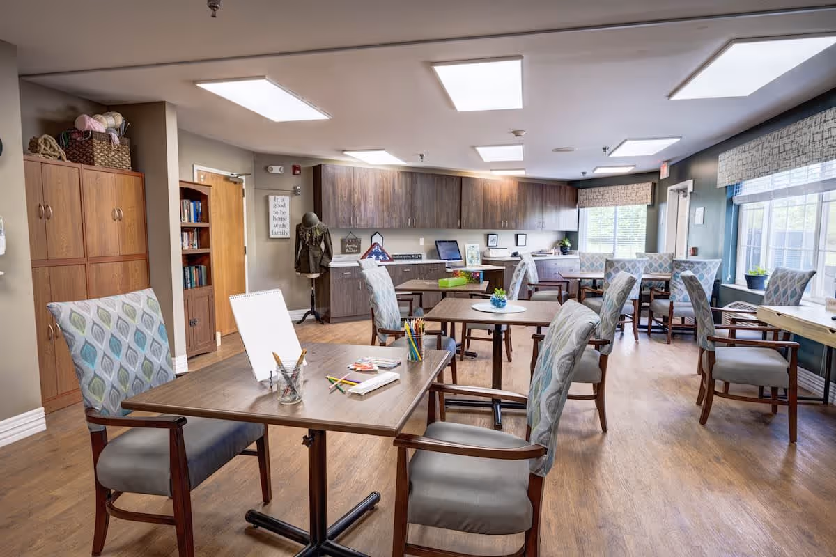 A bright and spacious activity room in a senior living facility with several tables and cushioned chairs arranged for group activities. The room features wooden flooring, large windows with patterned valances, and overhead fluorescent lighting. There are cabinets and shelves along the back wall, some with books and decorative items. Art supplies and papers are placed on the tables, suggesting a creative or recreational use.
