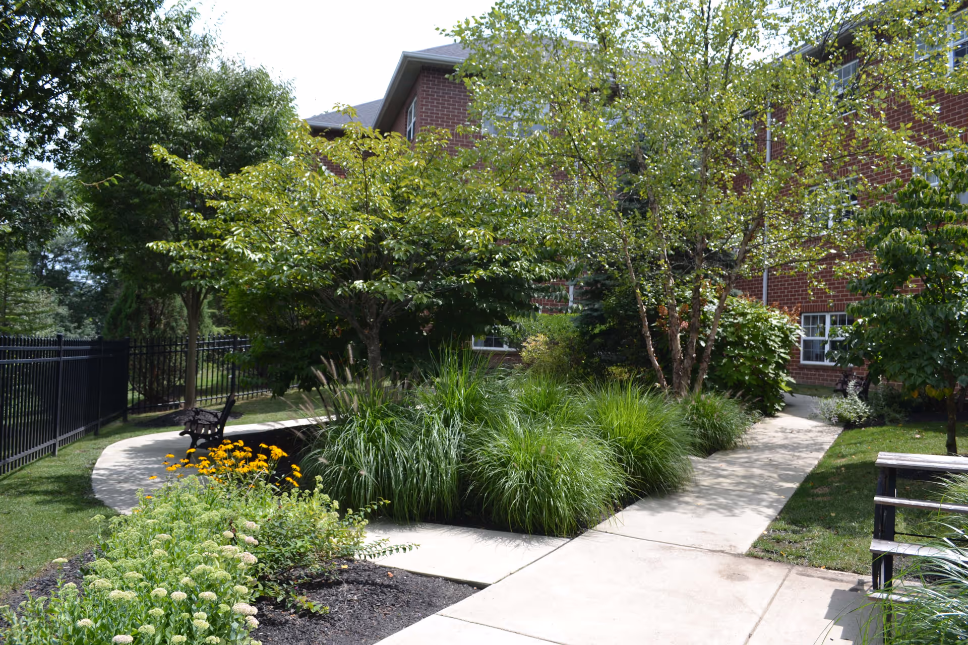 A landscaped outdoor garden area with a concrete pathway winding through green bushes, trees, and flowering plants. There is a black metal bench along the path and a red brick building in the background.
