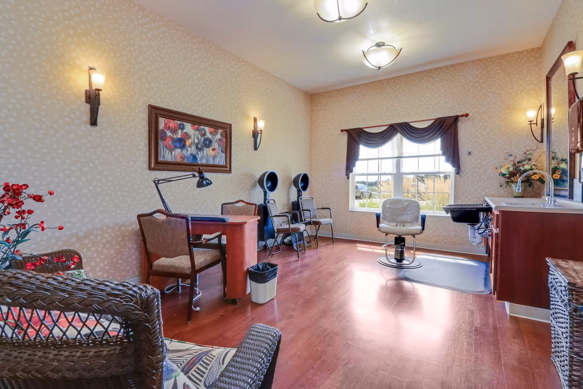 A well-lit room with wooden flooring featuring a salon chair in front of a large window with dark curtains. The room includes a desk with a lamp and chair, two hair drying chairs, a sink with a countertop and mirror, and wicker furniture with floral cushions. The walls have a light patterned wallpaper and are decorated with a framed floral painting and wall sconces.